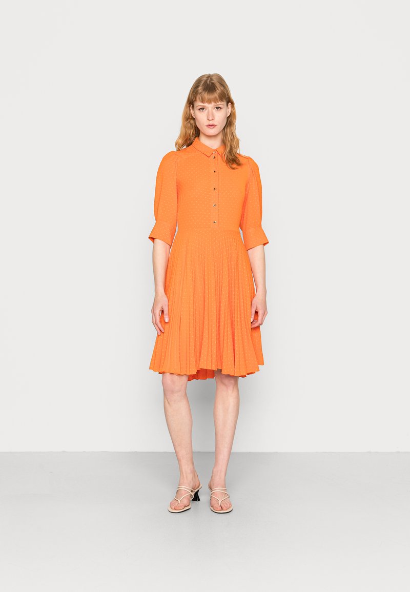 Vestido de manga curta, com colarinho, em laranja vibrante, com fecho de botão e uma saia fluída e plissada. Textura suave com detalhes em padrão de bolinhas.