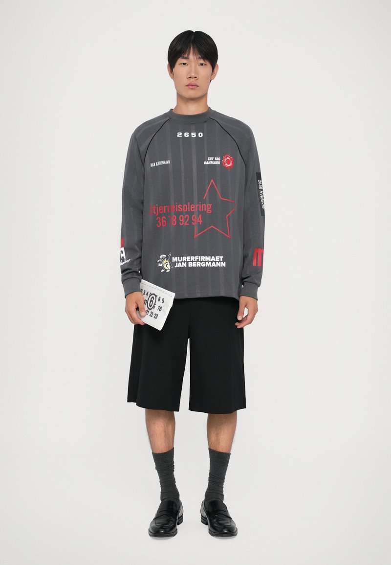 Long-sleeve gray top with red graphics, black shorts, gray socks, and black loafers. Accessories include a white rectangular bag.