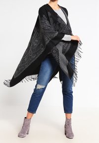 Poncho à motifs noir et gris avec des bords frangés, fabriqué dans un matériau doux. Porté par-dessus un haut gris et associé à un jean bleu usé.