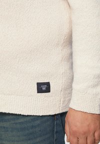 Cremfarbener Strickpullover mit strukturierter Oberfläche und gerippten Bündchen. Verfügt über ein schwarzes Etikett mit weißem Text in der unteren linken Ecke.
