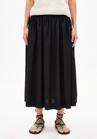 Schwarzer Maxikleid mit gerafftem Bund, aus leichtem Stoff. Kombiniert mit flachen Sandalen mit schwarzen Riemen und grünen Akzenten.