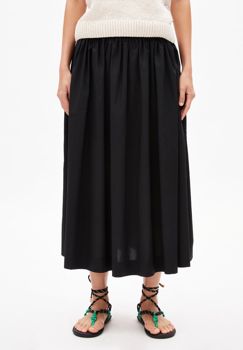 Schwarzer Maxikleid mit gerafftem Bund, aus leichtem Stoff. Kombiniert mit flachen Sandalen mit schwarzen Riemen und grünen Akzenten.