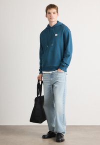 Mėlyna džemperis su raišteliais, priekinė kišenė, nešiojamas ant balto marškinėlio. Šviesiai skalbti plačio kojų džinsai ir juodi batai. Juoda rankinė.