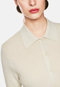 Cremfarbenes, geripptes Strickblusenoberteil mit klassischem Kragen und Knopfverschluss vorne. Der Stoff hat eine weiche, strukturierte Oberfläche mit dezentem Glanz.