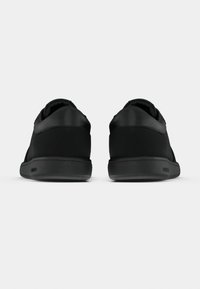 Ζεύγος μαύρων χαμηλών αθλητικών παπουτσιών εμφανίζεται από πίσω σε απλό λευκό φόντο, τονίζοντας τον ομαλό σχεδιασμό της φτέρνας και τις επίπεδες σόλες.