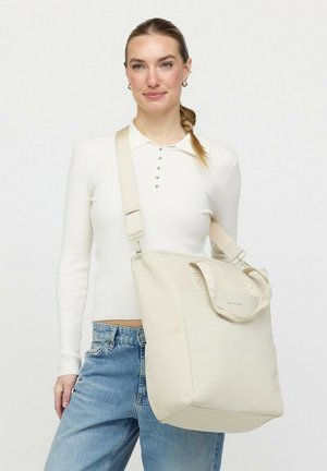 Vrouw draagt een witte top met lange mouwen en een spijkerbroek, en heeft een grote schoudertas in roomkleur met verstelbare band.