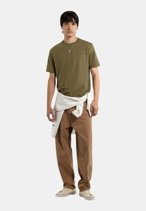 Giovane uomo che indossa una maglietta verde oliva, pantaloni marroni, scarpe da ginnastica bianche, una collana e un maglione bianco legato in vita.