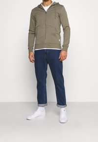 Sudadera verde oliva con cremallera y bolsillo delantero, llevada sobre una camiseta blanca. Combinada con jeans azul oscuro remangados y zapatillas blancas.