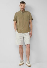 Olivgrünes Kurzarmhemd mit Brusttasche, kombiniert mit hellen strukturierten Shorts und beigen Sneakers mit braunen Akzenten.