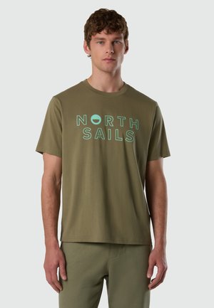 Joven con camiseta verde oliva de North Sails y pantalones a juego, posando frente a un fondo liso gris claro.