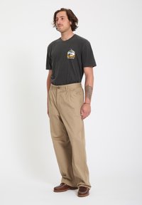 Homme aux cheveux mi-longs portant un t-shirt noir avec un motif graphique, un pantalon beige à jambes larges, des chaussures marron, et un tatouage sur l'avant-bras gauche, debout devant un fond blanc.