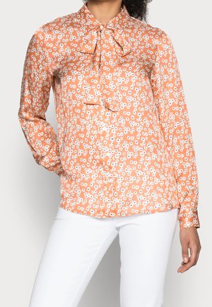 Blouse florale orange avec un nœud à la collerette, manches longues, texture lisse et motif de petites fleurs blanches avec des centres noirs.