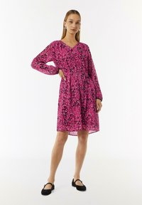 Rotes Kleid mit langen Ärmeln, V-Ausschnitt und tailliertem Schnitt. Es zeichnet sich durch ein kräftiges abstraktes Muster in Schwarz mit Farbtupfern in Pink aus.
