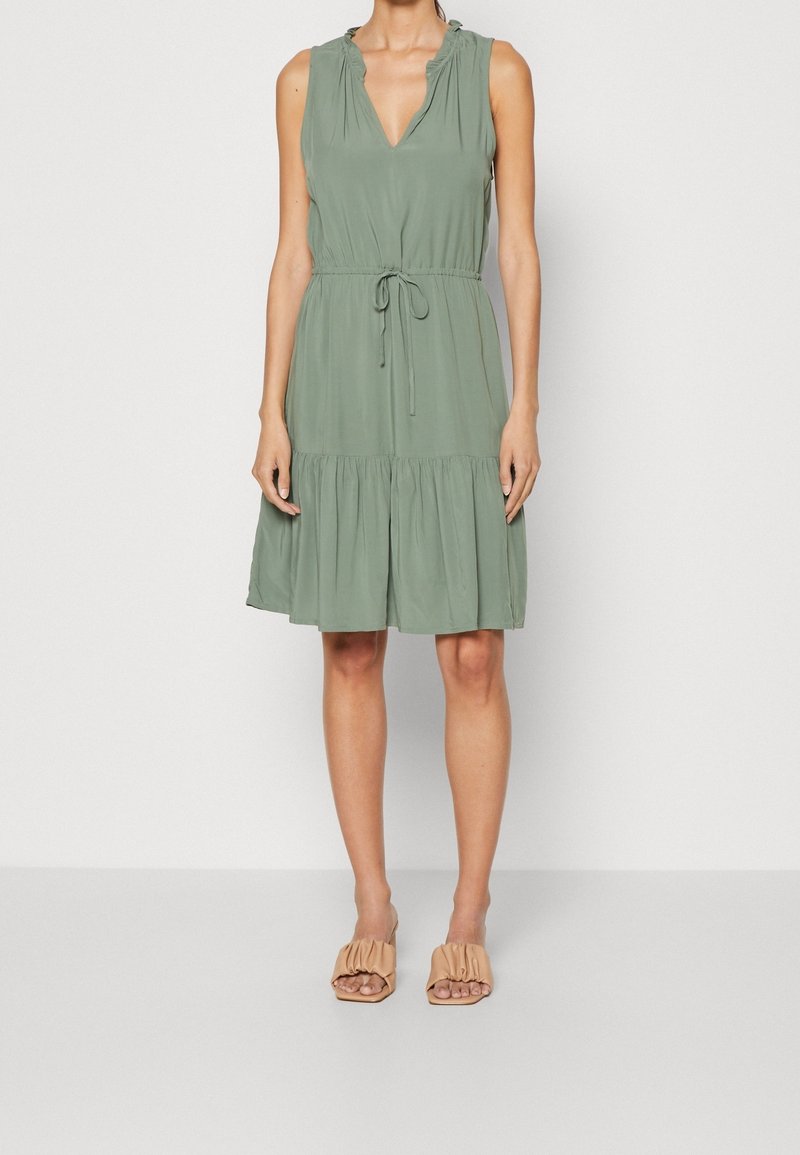 Femme portant une robe sans manches verte sauge longueur genoux avec taille resserrée par un cordon et des talons ouverts beige froncés, debout devant un fond blanc uni.