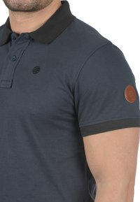 Námořnická polokošile vyrobená z texturového materiálu, s černým límcem, logem na hrudi a koženým štítkem na rukávu. Krátké rukávy.