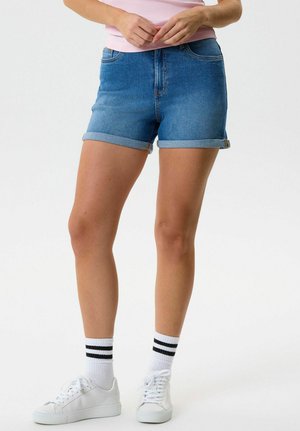 Jalad, mis kannavad ülesrullitud teksapükse, valgeid tossusid ja valgeid sokke kahe musta triibuga, käed kergelt talje juures kokku pandud.