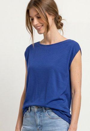 Junge Frau mit hellbraunen Haaren zu einem Dutt gebunden, trägt ein blaues ärmelloses Top und hellblaue Jeans, lächelt und schaut nach unten.