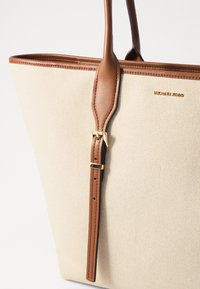 Borsa tote in tela beige con dettagli in pelle marrone, design strutturato, accessori in metallo dorato e dettaglio della tracolla regolabile. Logo esposto in modo prominente.