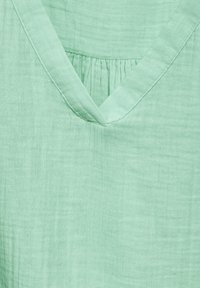 Top verde menta hecho de tela ligera que presenta un escote en V y un sutil detalle fruncido, con un acabado suave y texturizado.
