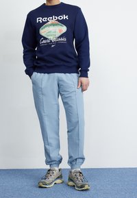 Mörkblå Reebok sweatshirt med grafiskt tryck, ljusblå strukturerade byxor och grå sneakers med gula accenter. Avslappnad passform, ribbade mudd.