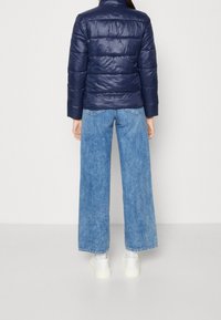 Granatowa kurtka puchowa z poziomym pikowaniem, w połączeniu z jasnoniebieskimi spodniami typu wide-leg i białymi sneakersami. Gładka faktura i wysoki kołnierz.