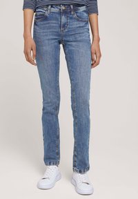 Vaqueros de mezclilla azul ajustados con un lavado claro, cinco bolsillos y detalles de costura. Combinados con zapatillas blancas.