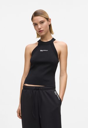 Femme portant un débardeur noir sans manches style dos nu avec le logo "Karl Lagerfeld Jeans" et un pantalon noir à cordon avec logo sur la poche.