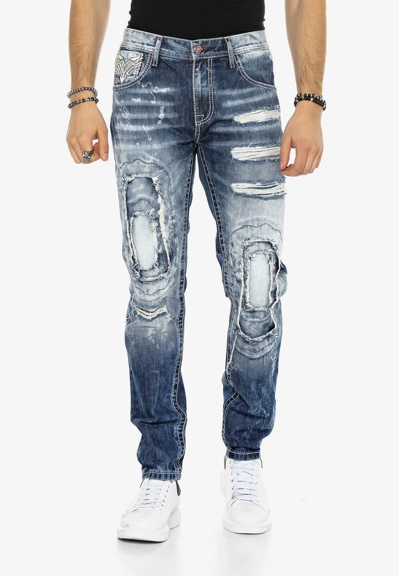 Abgenutzte blaue Jeans mit mehreren großen Rissen und ausgewaschenen Patches. Verfügt über ein Fünf-Taschen-Design und kontrastierende Nähte.