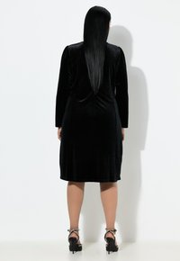 Czarna aksamitna sukienka z długimi rękawami, długość do kolan i gładka faktura. Masywne czarne sandały na obcasie z perłowymi akcentami.