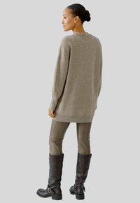 Beiger Pullover mit langen Ärmeln, gerippten Bündchen und Saum. Kombiniert mit eng anliegenden, glänzenden grünen Hosen und knielangen schwarzen Lederstiefeln.