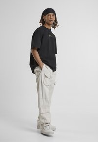 Svart oversized T-shirt med guldlogo; kombinerad med krämfärgade cargobyxor och ljusgrå sneakers. Slät textur, avslappnad passform, funktionella fickor.