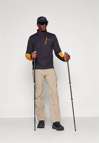 Vyras dėvi juodą striukę su oranžiniais akcentais, smėlio spalvos kelnes, juodus batus, kepurę ir akinius nuo saulės, laiko du žygiavimo lazdas priešais vientisą foną.