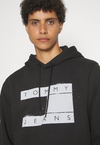 Felpa nera in cotone, con una grafica grigia e il testo "TOMMY JEANS". Include cordini e una vestibilità morbida.