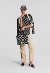 Borsa a tracolla in bianco e nero con manici doppi, abbinata a una camicia coordinata con motivo e pantaloni beige larghi. Il cappello presenta lo stesso design.