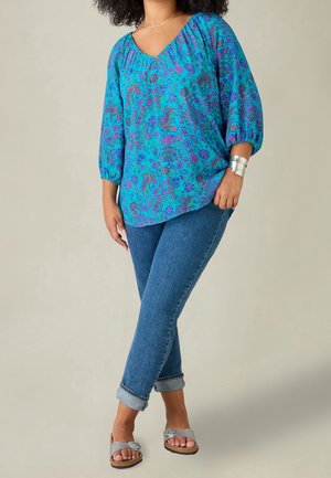 Woman wearing blue floral blouse with three-quarter sleeves, rolled-up blue jeans, silver bracelet, and silver sandals standing with legs crossed.