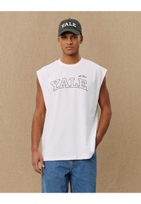 Camicetta senza maniche bianca con la scritta "YALE" in grassetto nero, abbinata a un cappellino verde scuro con lo stesso testo, su uno sfondo neutro.