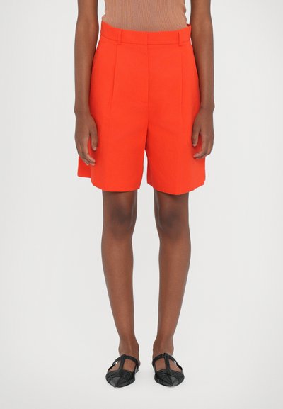 Shorts taillés orange avec des plis, taille haute, et poches latérales. Portés avec des ballerines noires en toile. Design simple, texture de tissu lisse.