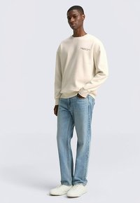 Lichtbeige sweatshirt met een ronde hals en een subtiele print op de borst, gecombineerd met lichtblauwe jeans en witte sneakers op een neutrale achtergrond.