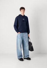 Felpa blu navy con tasca frontale e cappuccio con coulisse, abbinata a jeans a gamba larga blu chiaro. Borsa nera con accenti argentati. Calzature nere.