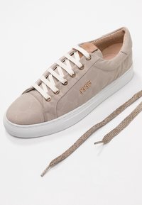 Baskets beige avec un design texturé, semelle en caoutchouc blanche, œillets dorés et un logo. Comprend un lacet métallique assorti.