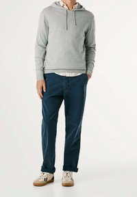 Felpa grigia con cappuccio e coulisse, indossata sopra una maglietta bianca, abbinata a pantaloni chinos blu navy e sneakers beige con dettagli scuri.