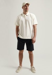 Hvit teksturert polo skjorte med tre knapper, kombinert med svarte shorts og beige sneakers, toppet med en lyse farget caps.
