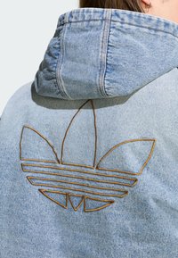 Világoskék farmerdzseki strukturált kapucnival. Barna, három szirmú és vízszintes vonalakkal díszített hímzett logó található a hátoldalon.