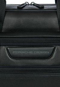 Sac en cuir noir avec une surface texturée, une fermeture éclair supérieure et un logo "Porsche Design" en relief très visible.