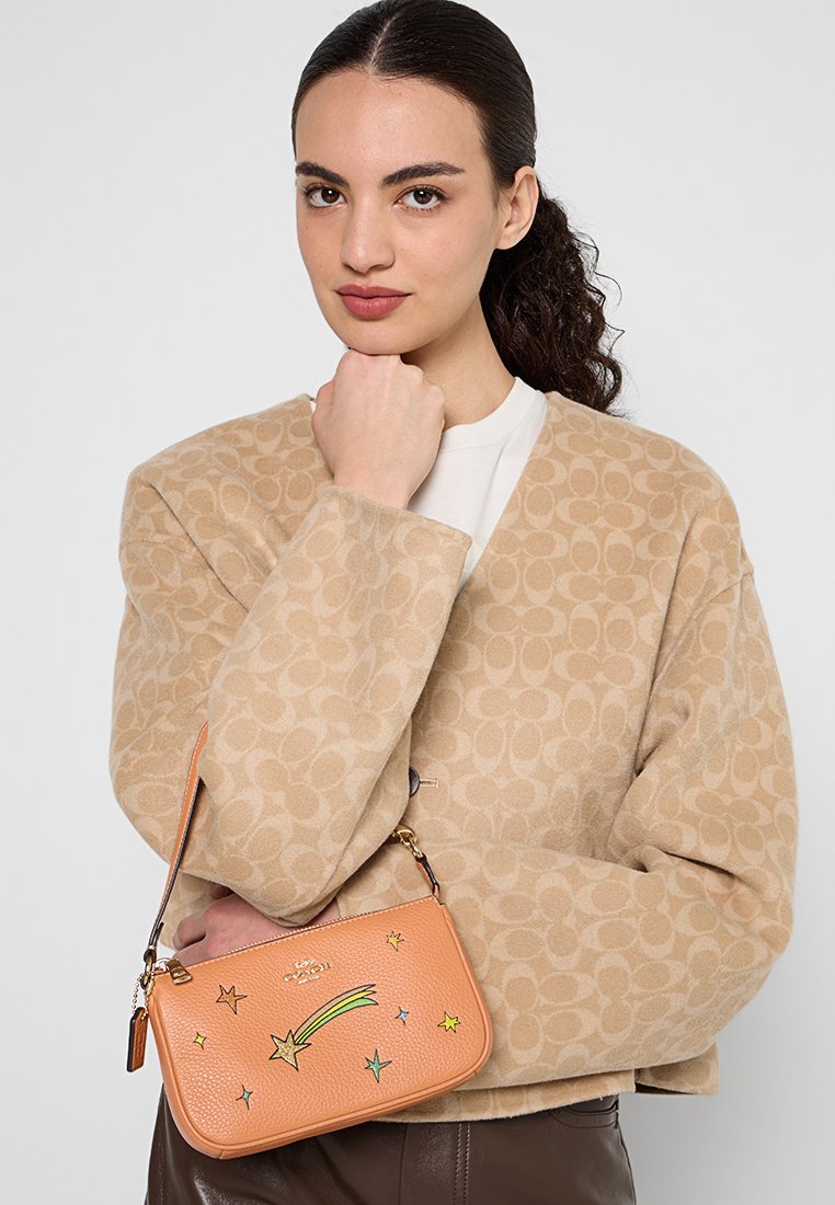 Vrouw met donker haar naar achteren getrokken, draagt een beige jas met patroon, houdt een oranje leren tas vast versierd met kleurrijke vallende sterren.