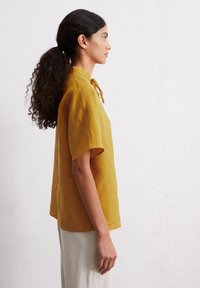 Short-sleeve mustard yellow blouse with a relaxed fit and tie neck detail, made of lightweight fabric, showcased from the side.