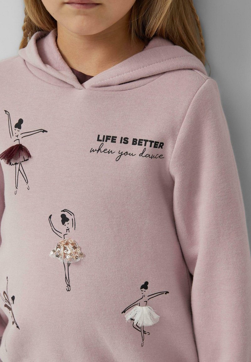 Dziecko w różowym hoodie z ilustracjami czarnych tancerek baletowych i cekinowymi spódnicami, tekst brzmi: "Życie jest lepsze, kiedy tańczysz."