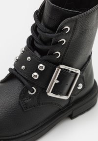 Botín negro de cuero hasta el tobillo con diseño de cordones, correa con hebilla metálica, tachuelas plateadas, superficie texturizada y suela plana.
