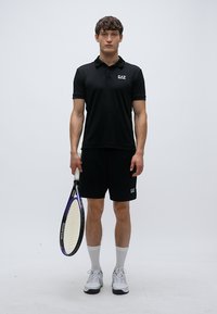 Čierny polo a šortky, ktoré ukazujú priliehavý strih. Materiál vyzerá ľahký. Hráč drží tenisovú raketu s fialovými a čiernymi detailmi.