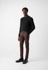 Μαύρος πλεκτός σκούφος, ελιάς τζάκετ με ψηλό γιακά, καφέ στενό παντελόνι και μαύρα παπούτσια με κορδόνια. Απλή σχεδίαση με ραβδωτή υφή και λεία φινιρίσματα.